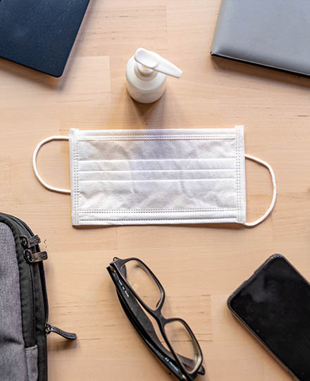 Desk containing glasses, laptop, laptop bag, mobile phone and a centered surgical mask and sanitiser